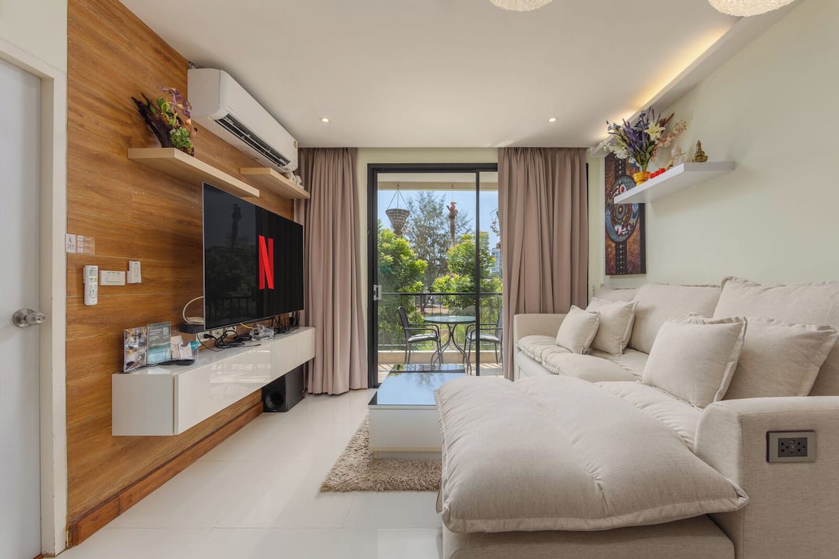 A modern living room with a flat-screen TV, a sofa, a coffee table, and a balcony with a view.
