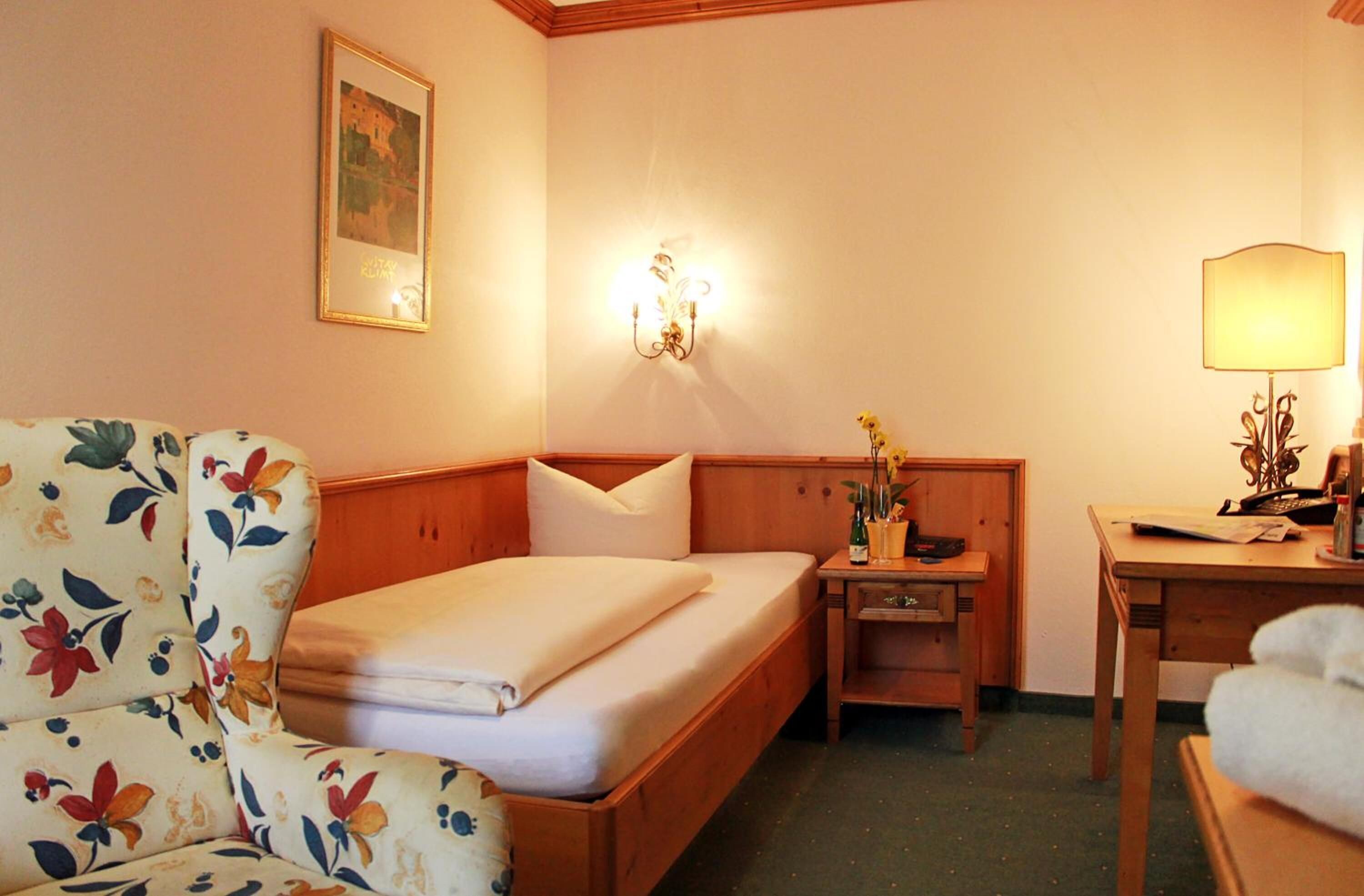 A hotel room with a bed, a floral armchair, a nightstand with a lamp, and a small table with a telephone.