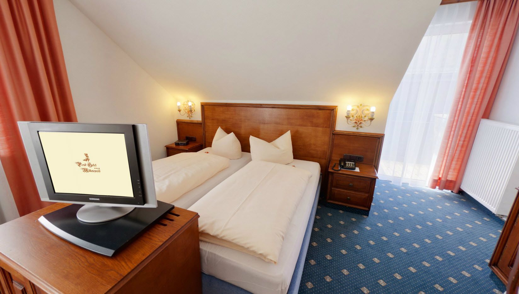 A hotel room with a bed, a desk with a computer, a nightstand, and a window with curtains.