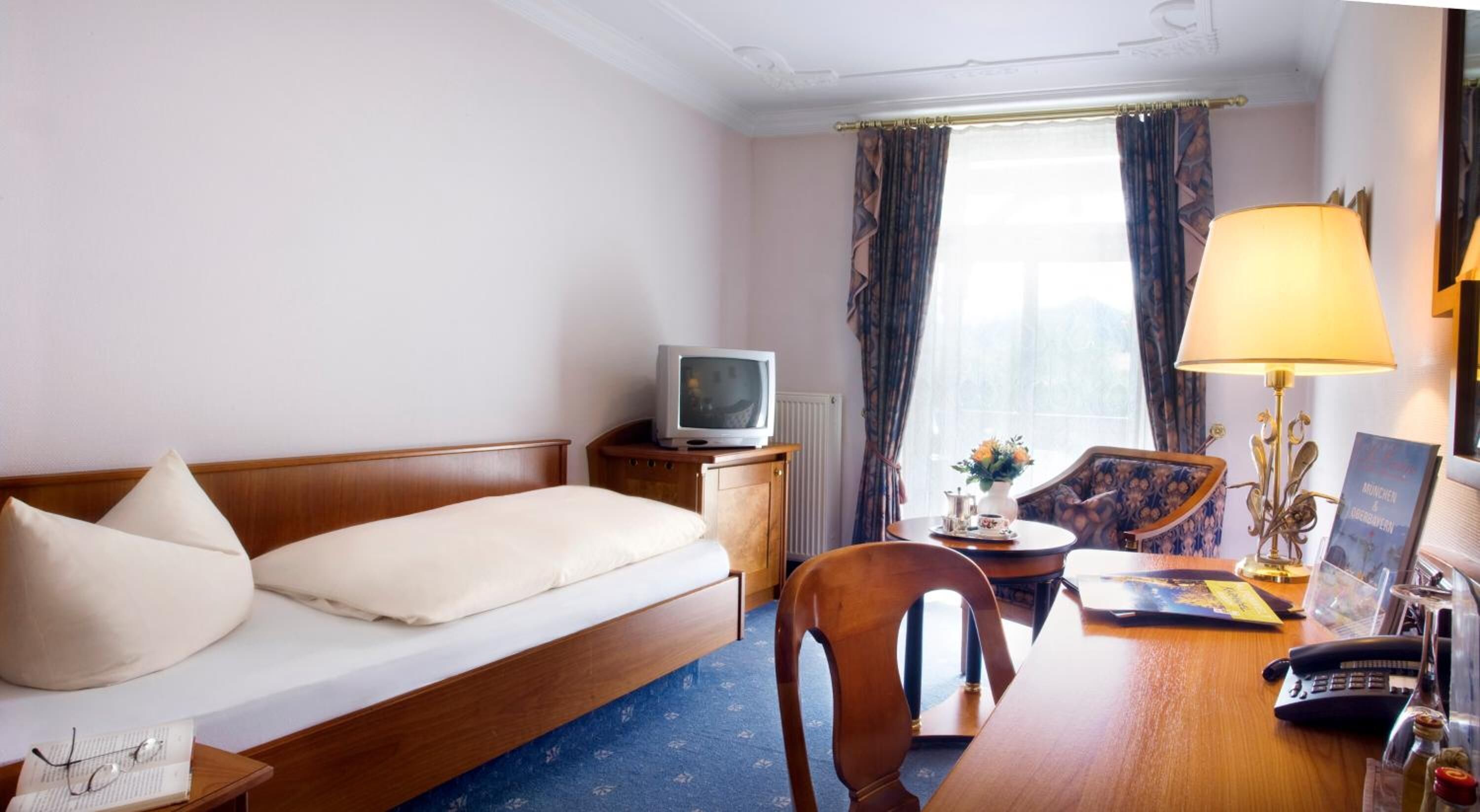 A hotel room with a bed, a desk with a lamp, a chair, a television, and a window with curtains.