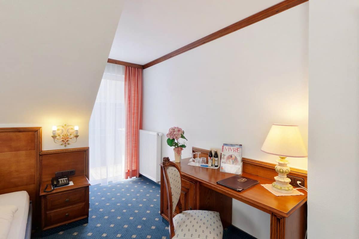 Habitación de hotel con cama, escritorio con lámpara, silla, mesita, teléfono, radiador, ventana con cortinas y una lámpara de pared.