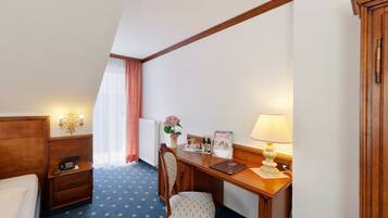 Habitación de hotel con cama, escritorio con lámpara, silla, mesita, teléfono, radiador, ventana con cortinas y una lámpara de pared.