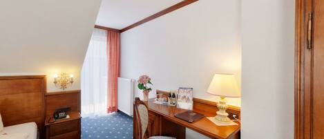 A hotel room with a bed, a desk with a lamp, a chair, a small table, a telephone, a radiator, a window with curtains, and a wall-mounted light fixture.