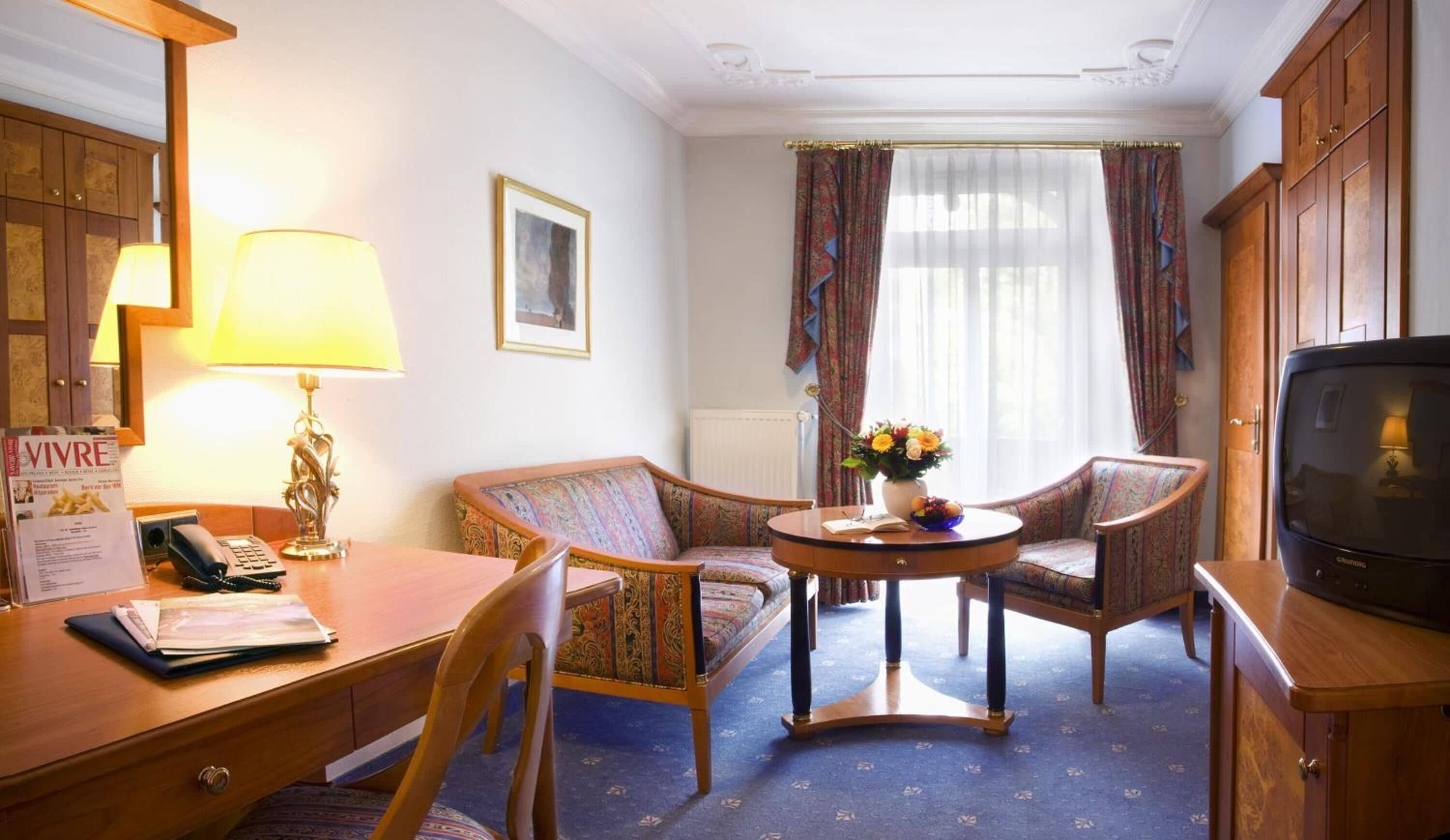 Ein Hotelzimmer mit einem Holztisch, zwei Sesseln, einem kleinen Tisch mit Blumenvase, einem Fernseher und einem Fenster mit Vorhängen.