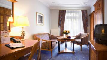 Ein Hotelzimmer mit einem Holztisch, zwei Sesseln, einem kleinen Tisch mit Blumenvase, einem Fernseher und einem Fenster mit Vorhängen.
