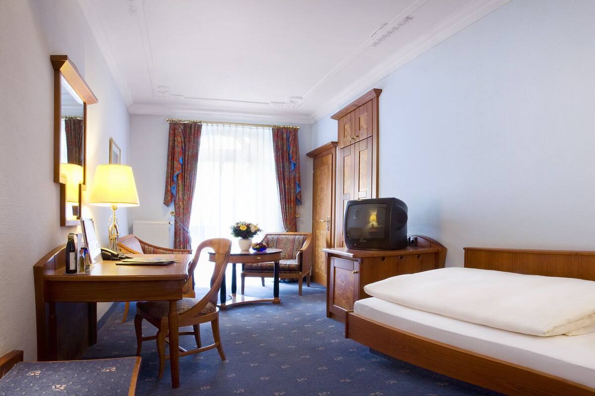 Habitación de hotel con una cama, un escritorio con lámpara, una silla, un televisor y una mesita con un florero.