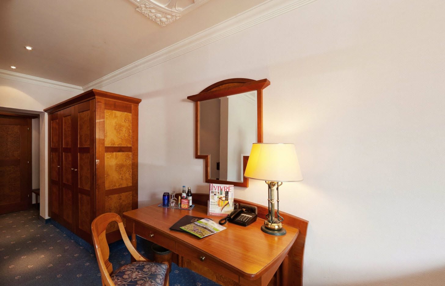 A hotel room with a wooden desk, a chair, a lamp, a mirror, and a minibar.