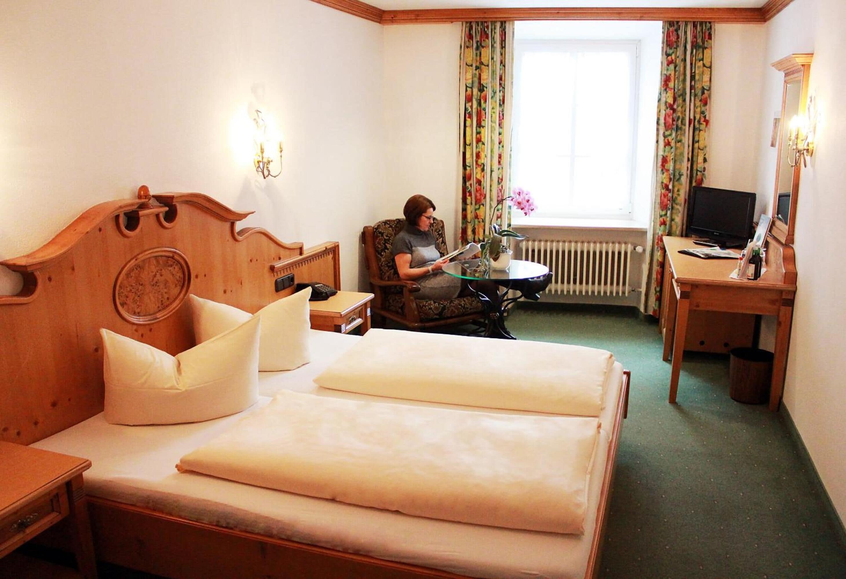 Habitación de hotel con una cama de madera, una silla, una mesita, un escritorio con una computadora y una ventana con cortinas.