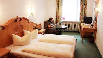 Habitación de hotel con una cama de madera, una silla, una mesita, un escritorio con una computadora y una ventana con cortinas.