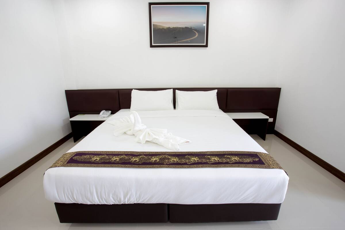 Una cama bien tendida con una colcha blanca y morada, dos almohadas blancas y una toalla blanca doblada sobre la cama.