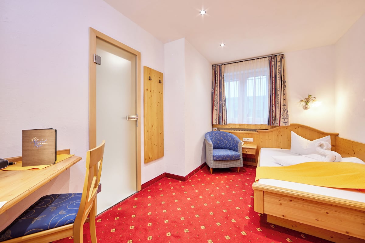A hotel room with a wooden desk, a chair, a bed with a yellow blanket, a blue patterned chair, a wooden wardrobe, and a window with patterned curtains.