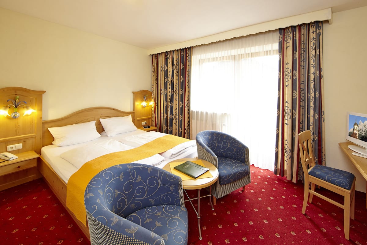 A hotel room with a bed, two armchairs, a small table, a desk, and a television.