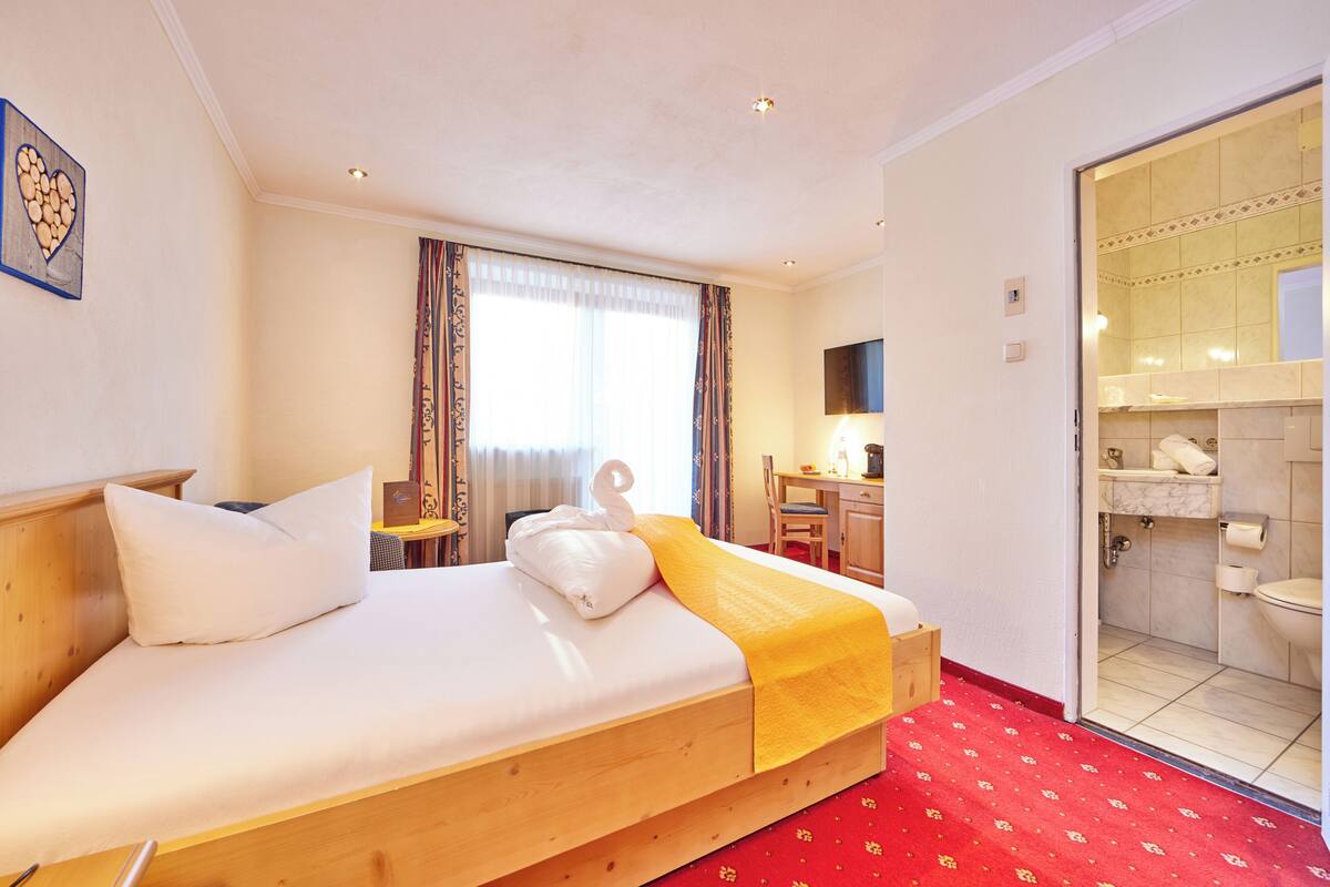 A hotel room with a wooden bed, a desk, a chair, a bathroom visible through an open door, and a red carpet.