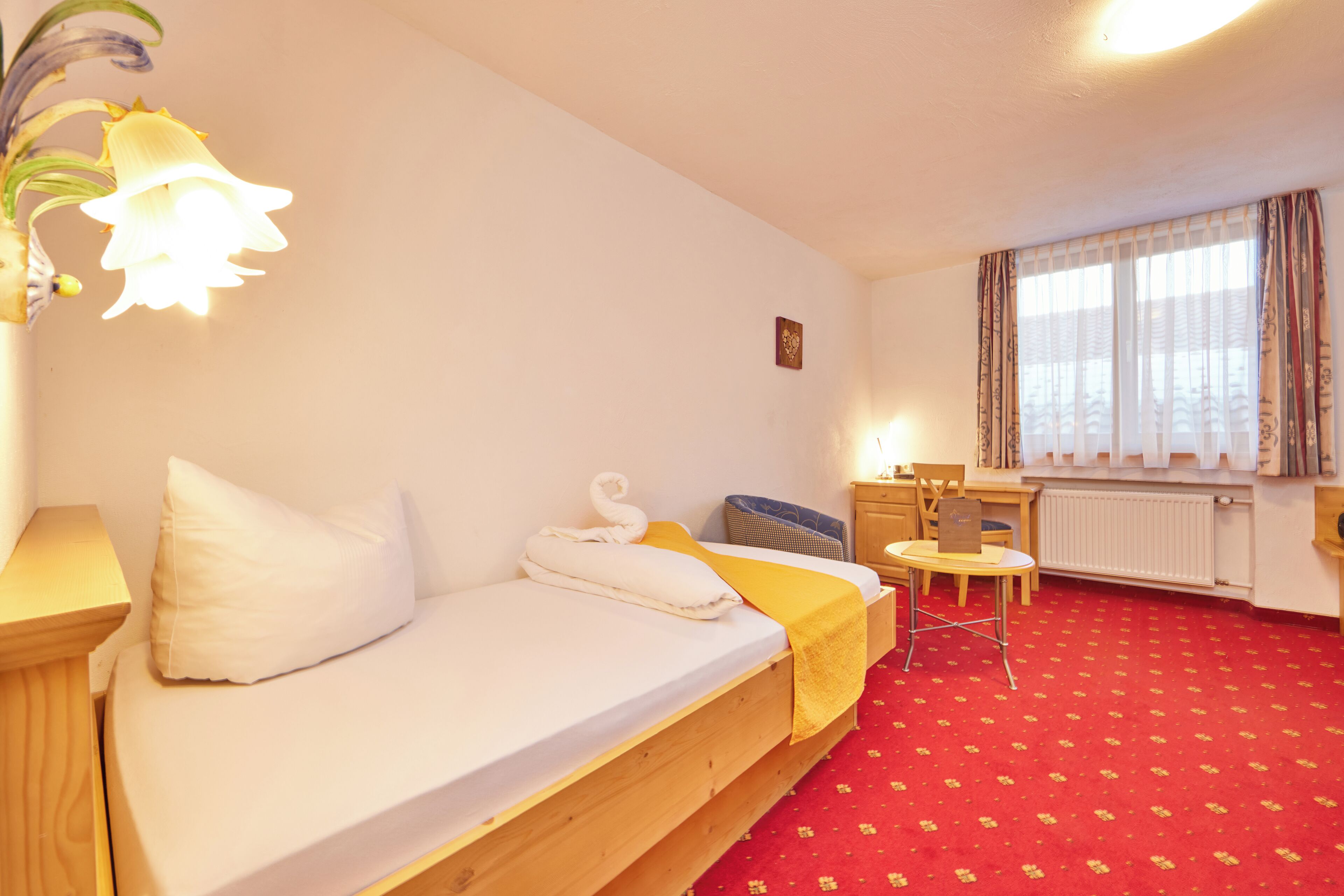 A room with a bed, a small table, a chair, a desk, and a window with curtains.