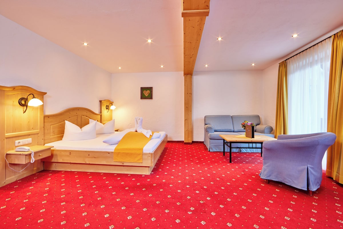 A hotel room with a wooden bed, a sofa, a chair, a small table, and a red carpet.