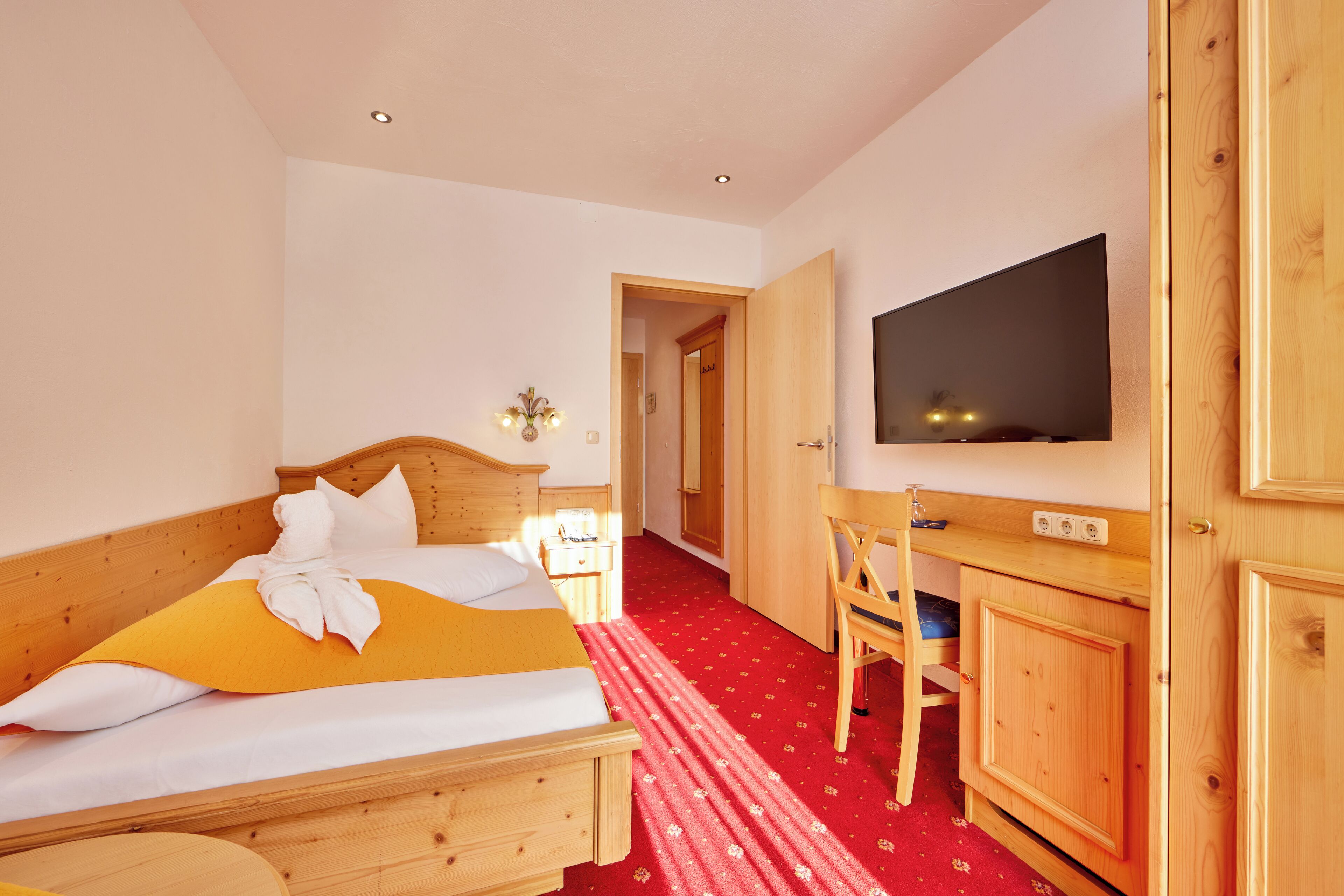 Habitación de hotel con una cama de madera, un escritorio con una silla, un televisor y una alfombra roja.