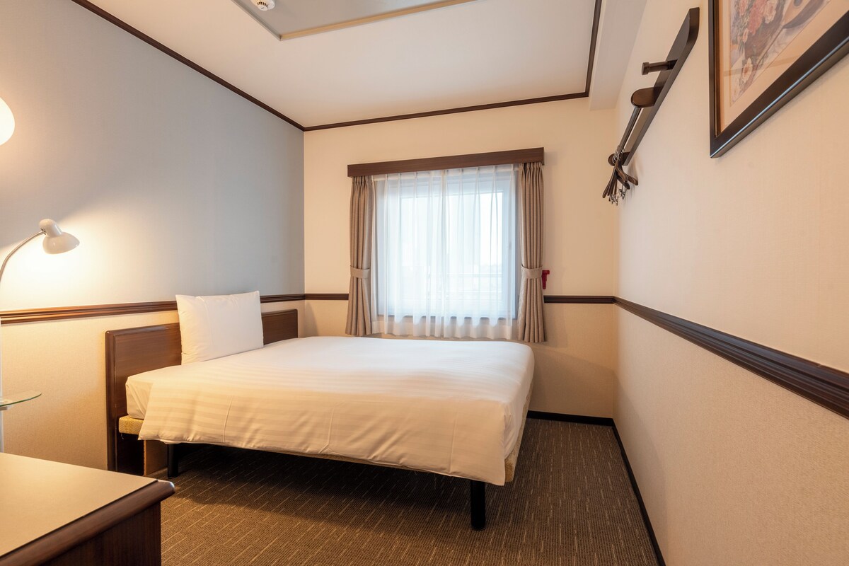 Et hotelværelse med seng, natbord, lampe og et vindue med gardiner.
