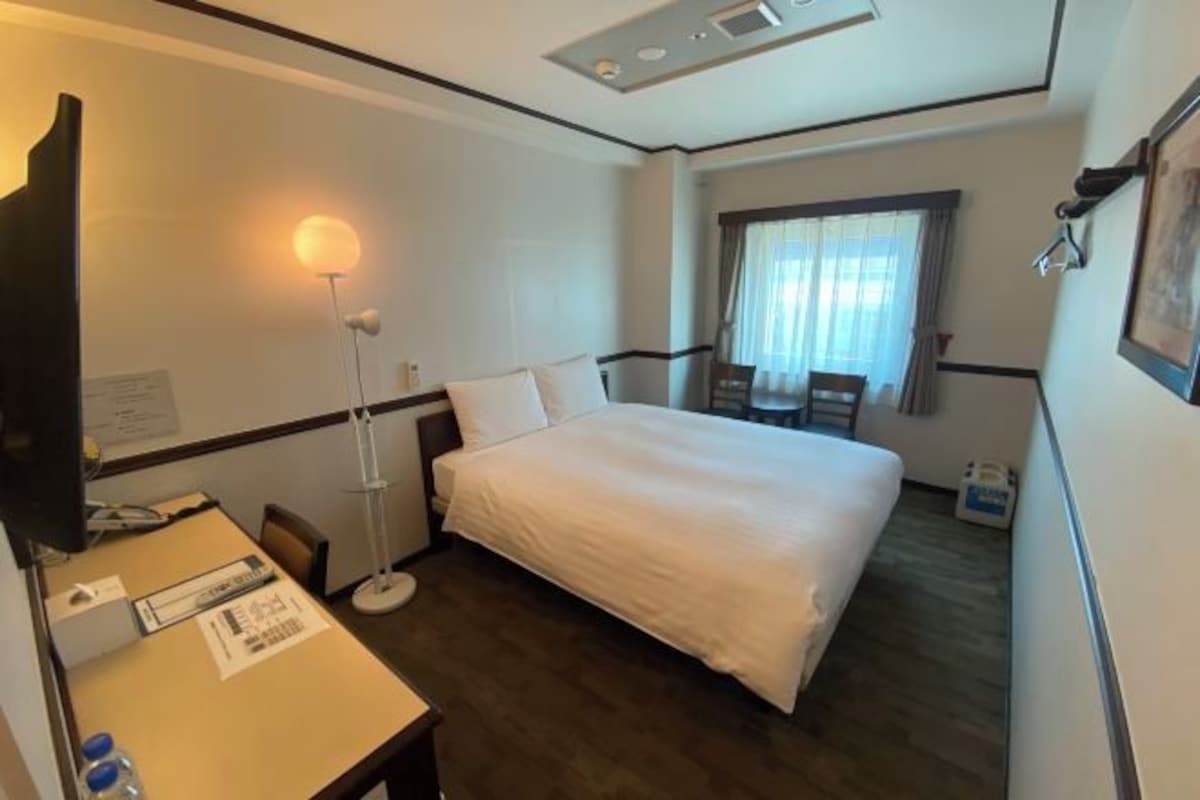 Quarto de hotel com cama, escrivaninha com cadeira, abajur e janela com cortinas.