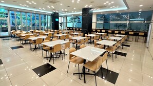 A spacious dining area with multiple tables and chairs, large windows offering views of the outdoors, and a modern ceiling design with recessed lighting.