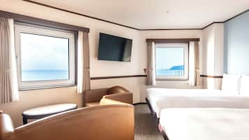 A hotel room with two beds, a flat-screen TV, a seating area, and large windows offering a view of the sea.