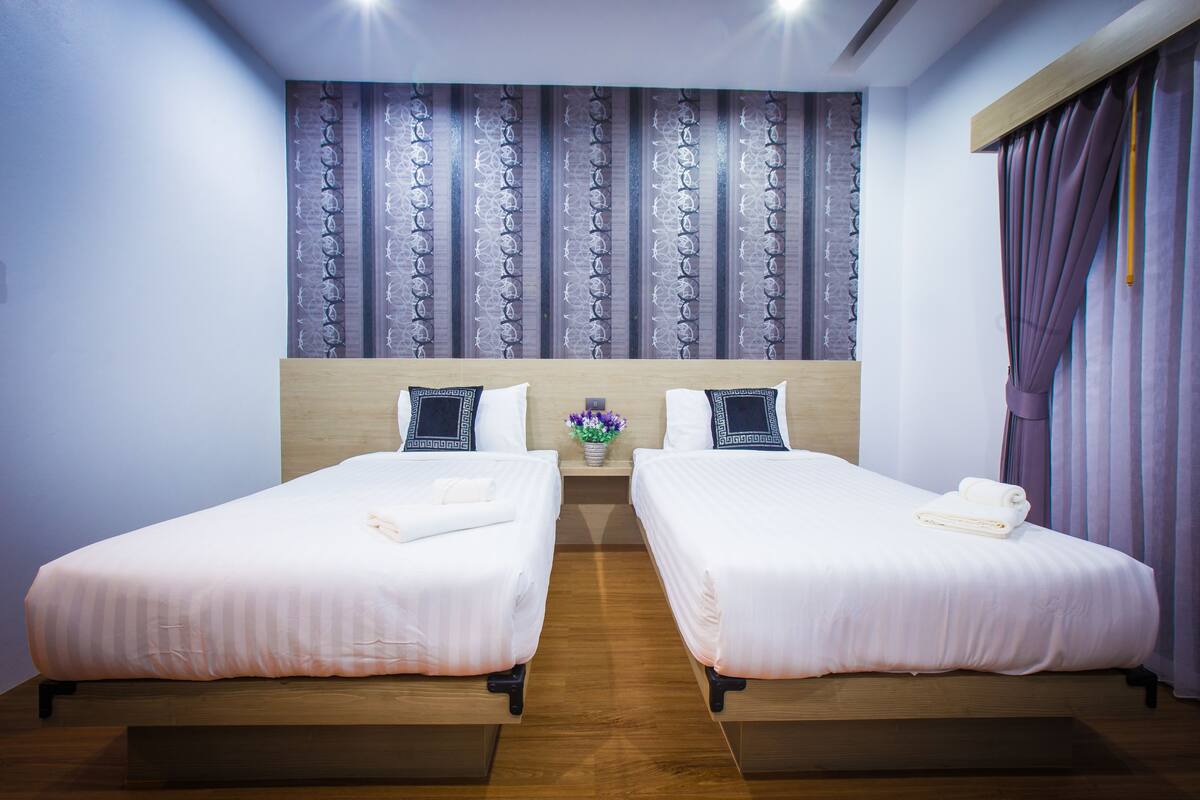 Habitación de hotel con dos camas individuales, cabeceros de madera, una mesita de noche con un florero y un ventanal con cortinas.