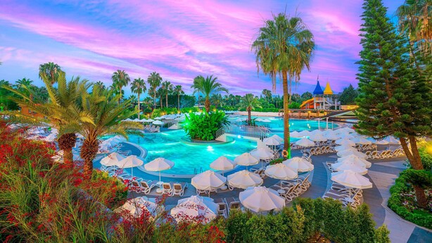 A resort pool area with lounge chairs and umbrellas, surrounded by palm trees and a colorful sky. - Arycanda Kirman Premium - Ultra All Inclusive (Alanya)
