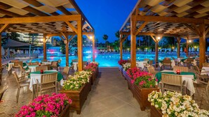 An outdoor dining area with tables and chairs, a pool in the background, and a wooden pergola structure. - Arycanda Kirman Premium - Ultra All Inclusive (Alanya)