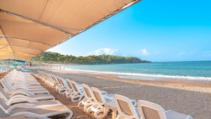 A beach with rows of white lounge chairs under a canopy, a sandy shore, and a clear view of the sea and distant land. - Arycanda Kirman Premium - Ultra All Inclusive (Alanya)