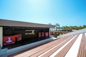 A modern hotel lobby with a stage area for events, a wooden deck, and palm trees in the background. - Sidera Kirman Premium - Ultra All Inclusive (Alanya)