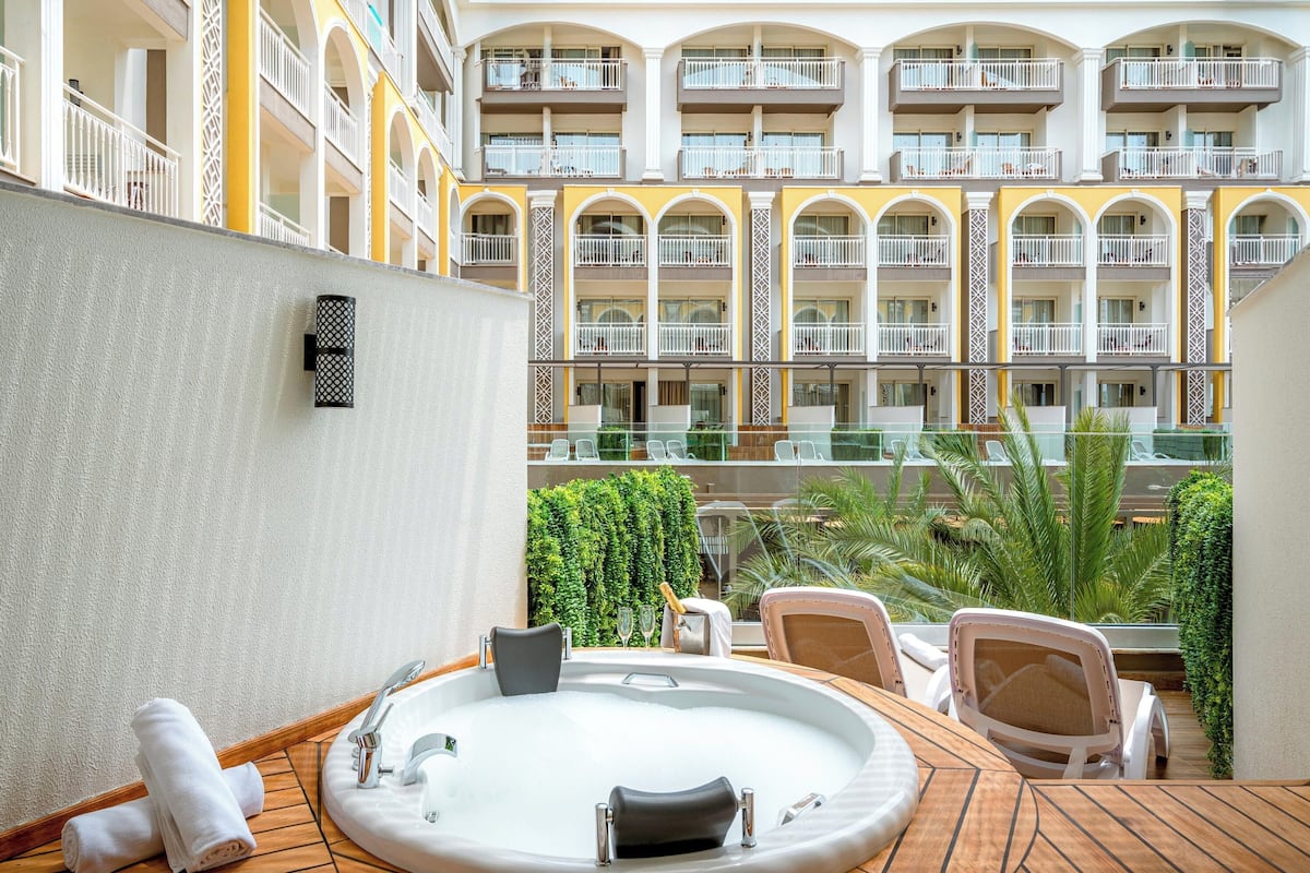 Un balcon d’hôtel avec un jacuzzi, des chaises longues et une vue sur les balcons intérieurs de l’immeuble.