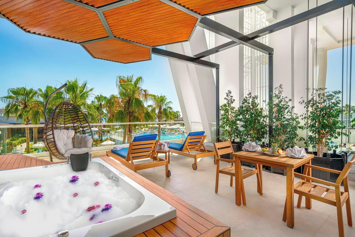 Une terrasse sur le toit avec un jacuzzi, des chaises longues en bois, une table avec des parasols et une vue sur des palmiers et une piscine.