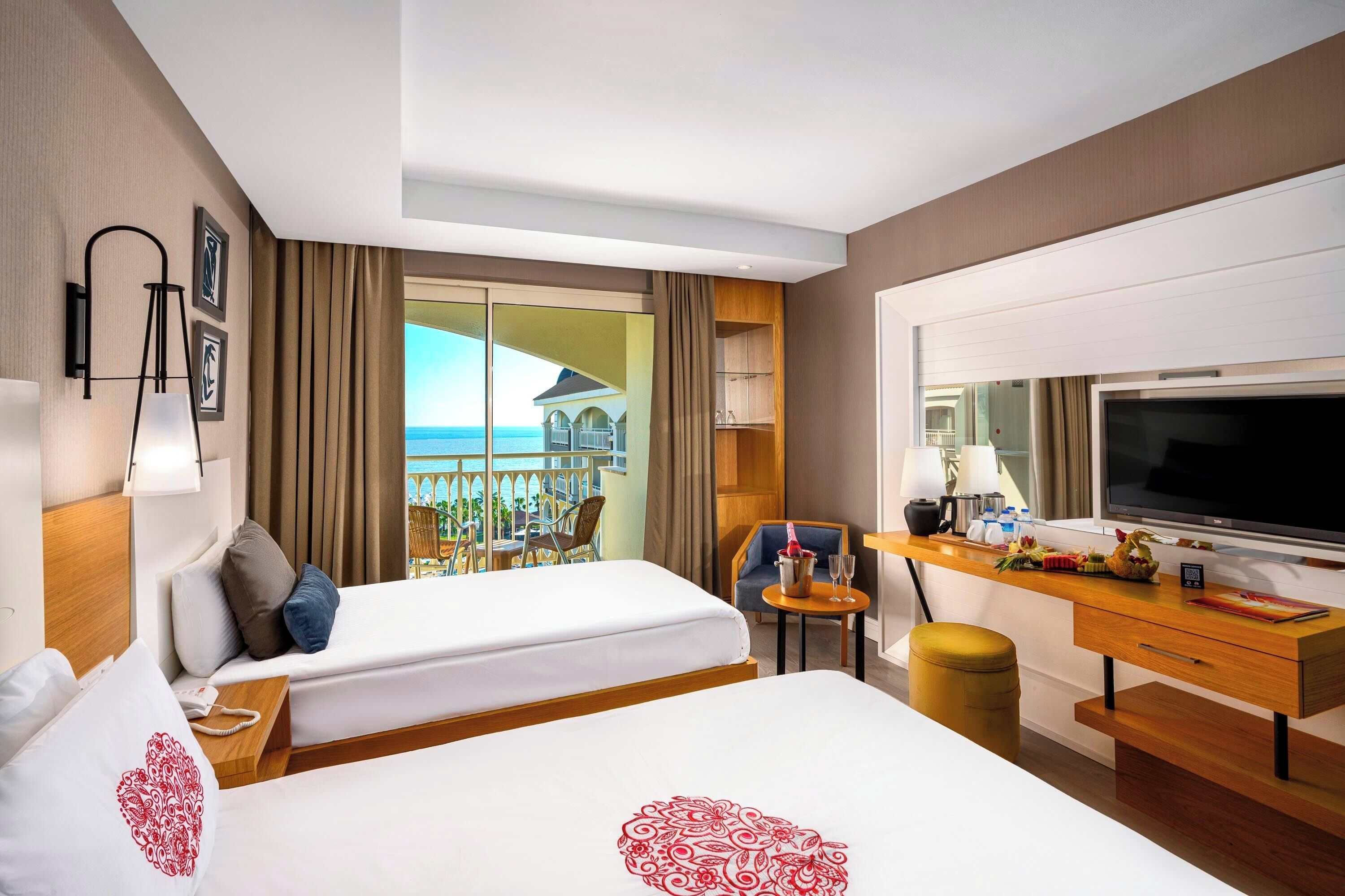 Habitación de hotel con dos camas, un escritorio con televisor, un balcón y vistas al mar, y una silla.