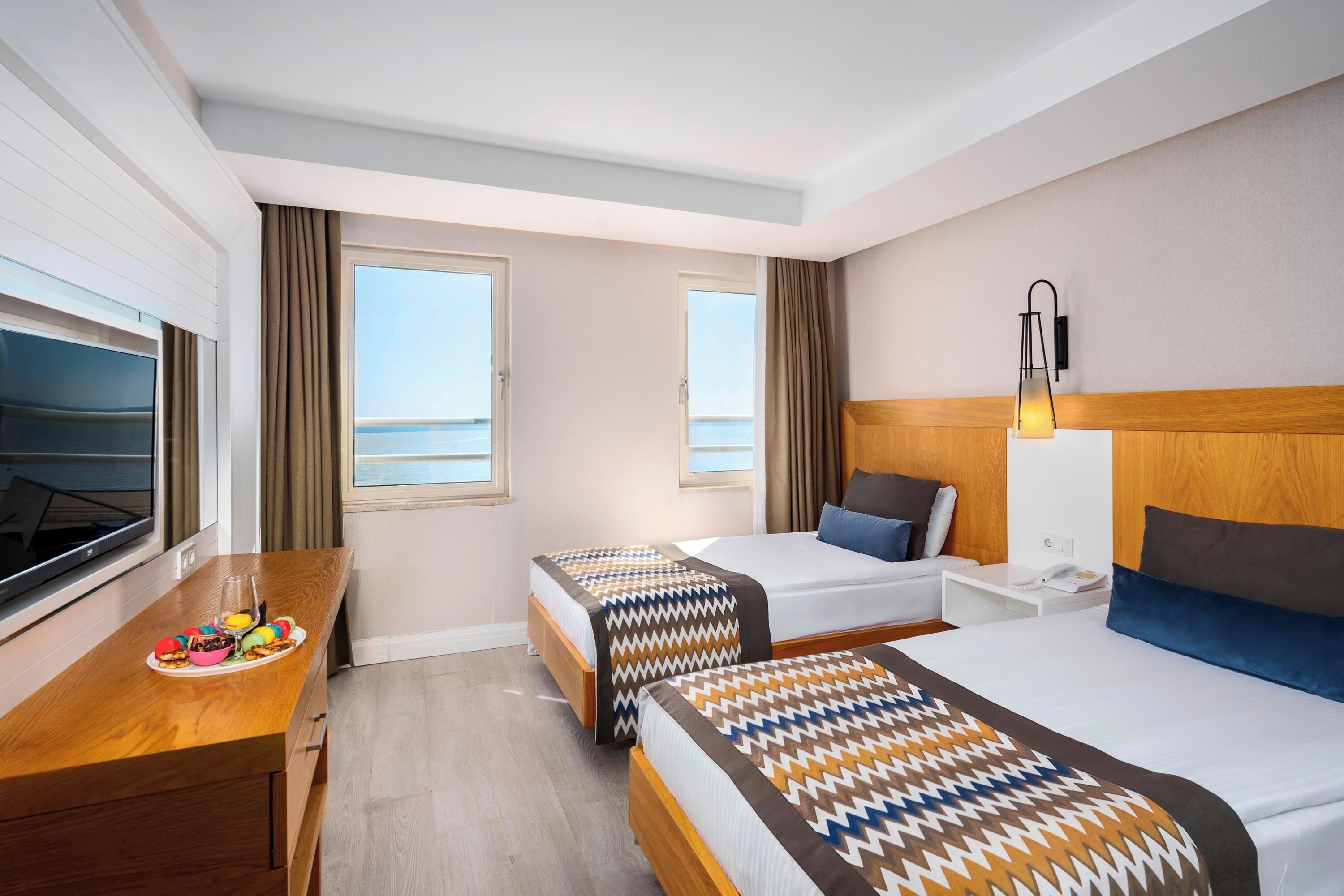 A hotel room with two beds, a wooden desk, a TV, and a view of the sea.