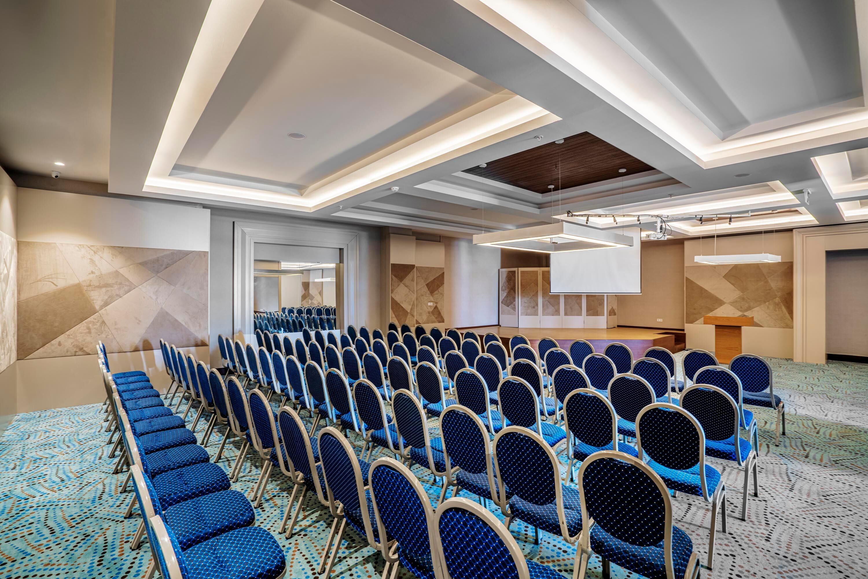 A conference room with rows of chairs, a podium, and a large screen.