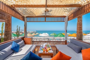 Un espacio de estar al aire libre cubierto con una pérgola de madera, asientos acolchados, una mesa con una bandeja de frutas y vistas a la playa y al océano.