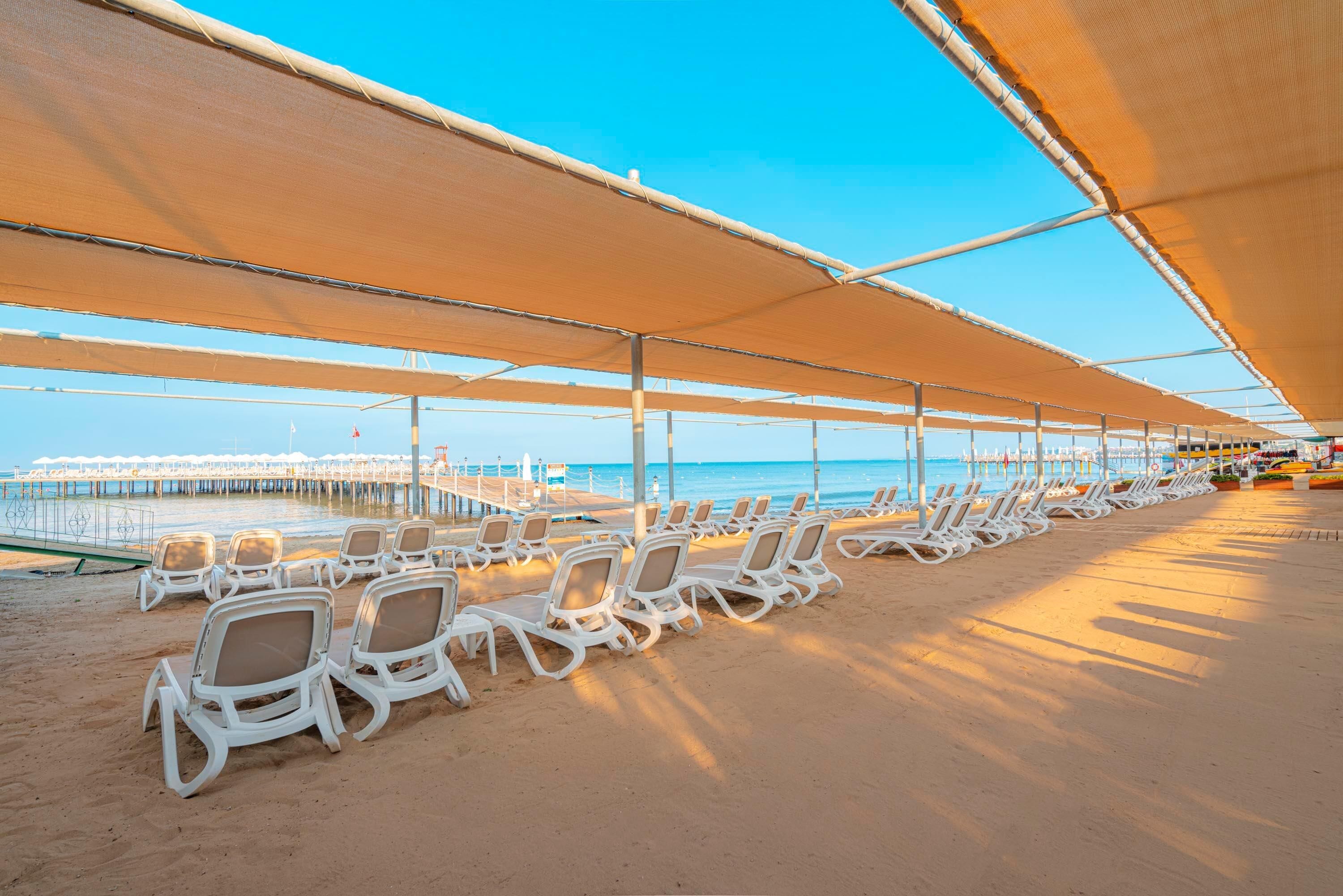 A beachside area with rows of white lounge chairs under a large canopy, a pier in the background, and a clear blue sky.