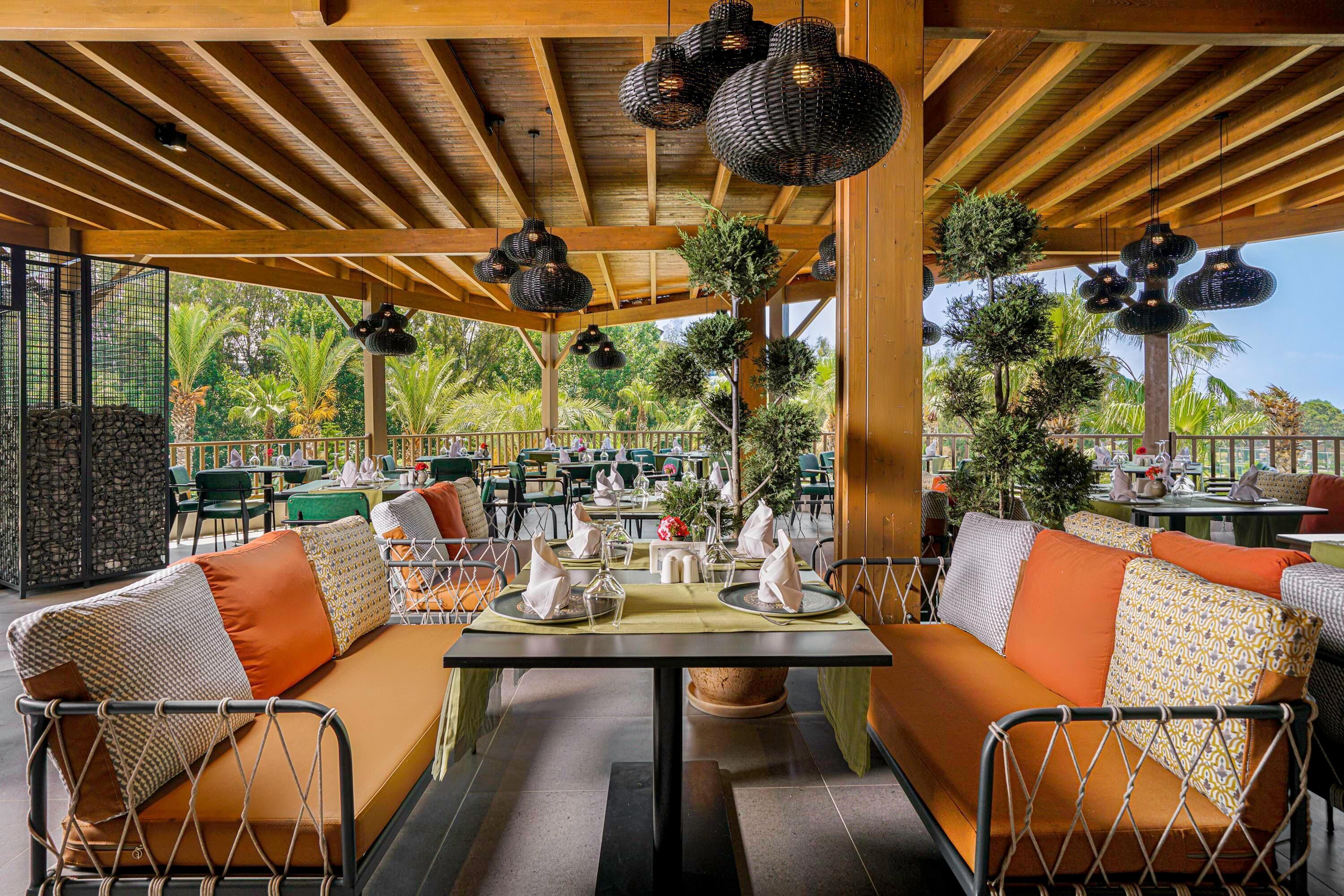 A covered outdoor dining area with wooden beams, wicker pendant lights, and green seating with orange cushions.