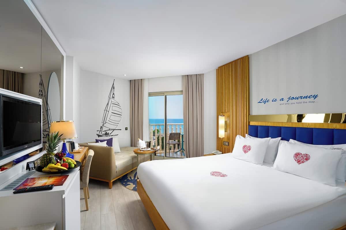 Een hotelkamer met een bed, een bureau met een televisie, een stoel, een kleine tafel met fruit en uitzicht op zee.