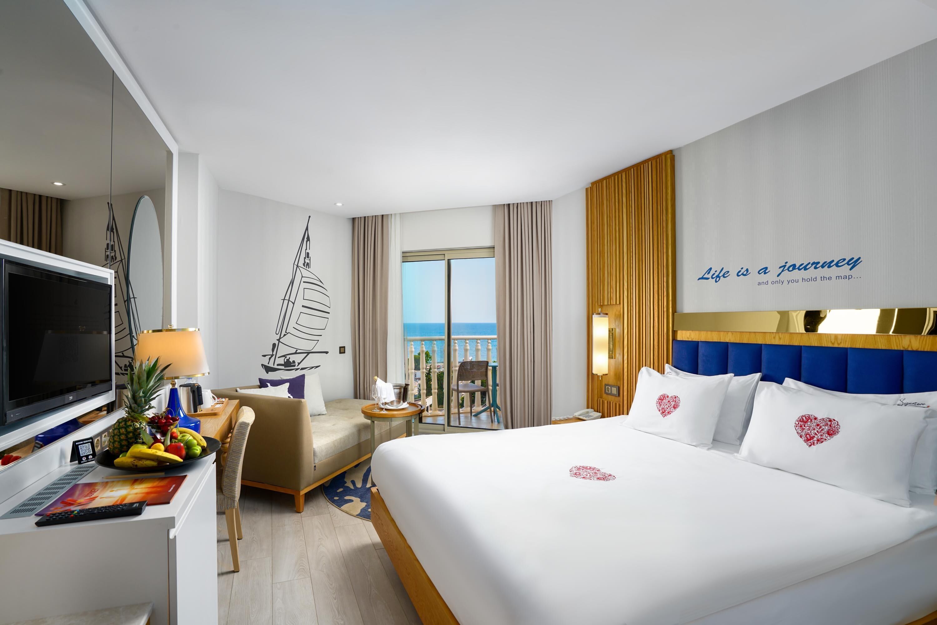 Ein Hotelzimmer mit Bett, Fernseher, Schreibtisch mit Lampe, Stuhl, Obstschale und Meerblick.