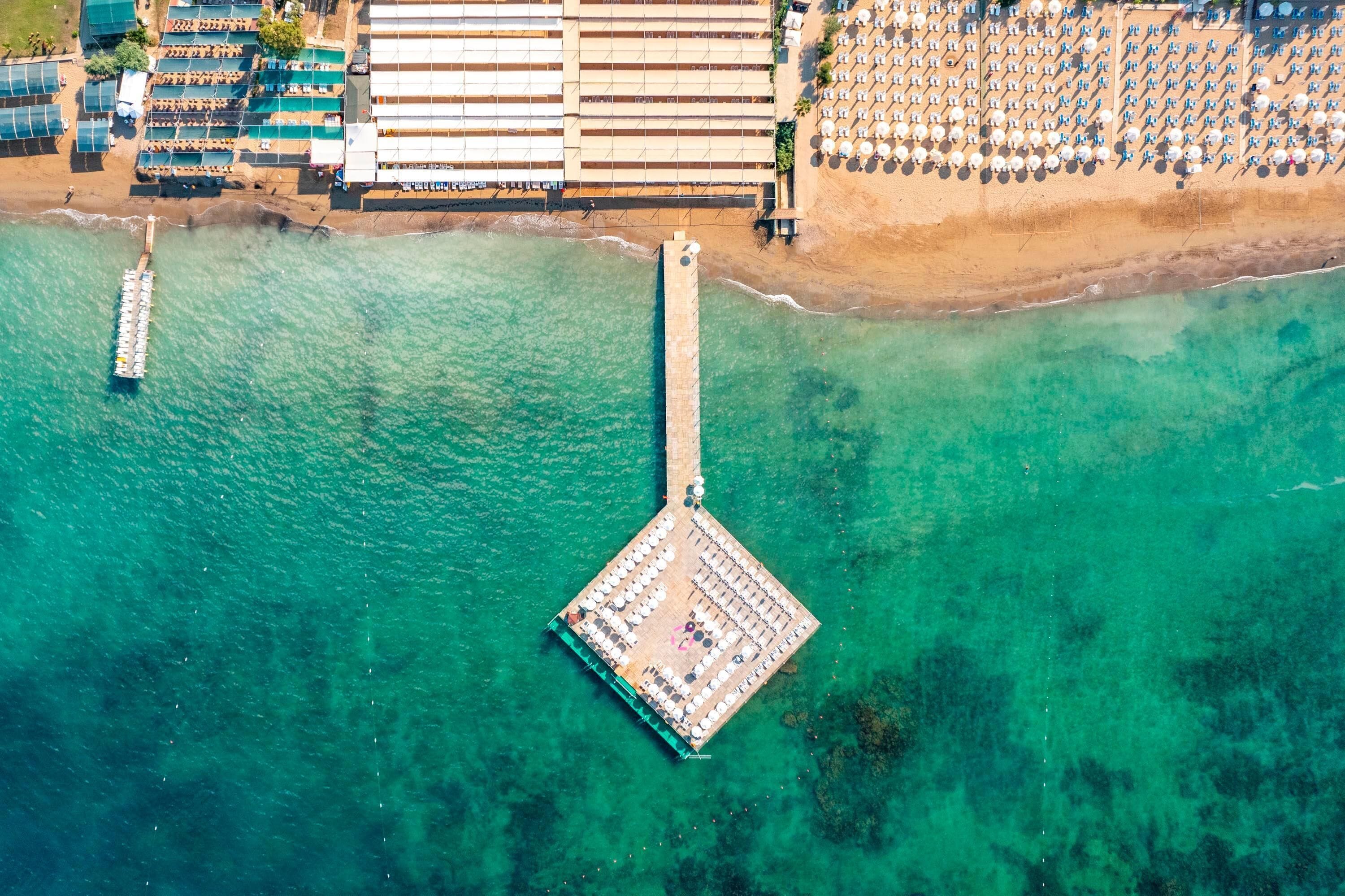 An aerial view of a coastal hotel with a pier, beach umbrellas, and a pool area.