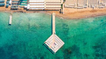 An aerial view of a coastal hotel with a pier, beach umbrellas, and a pool area.