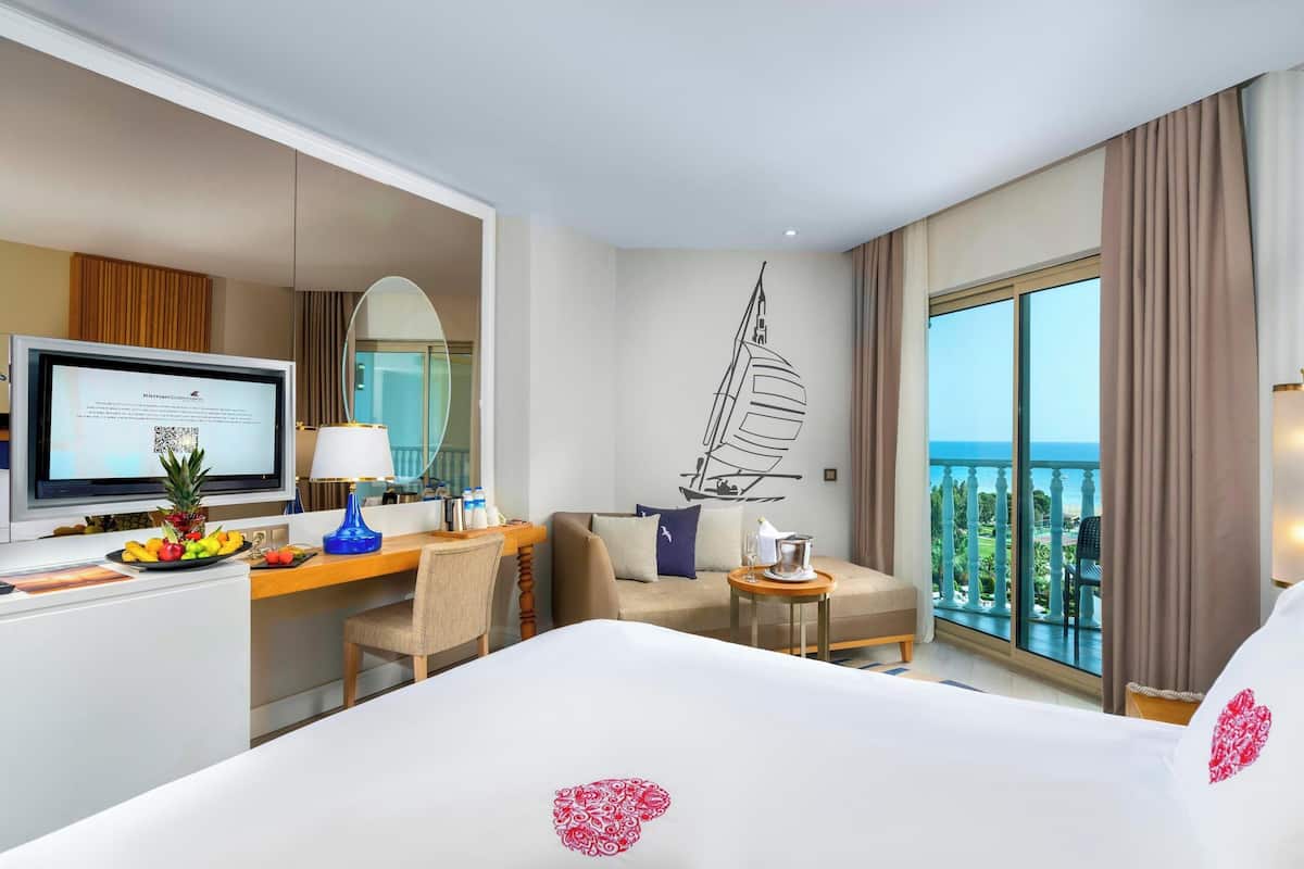 Hotelkamer met een bed, een bureau met een computer, een televisie, een fruitschaal, een stoel, een bank en een klein tafeltje met een lamp, plus een balkon met uitzicht.
