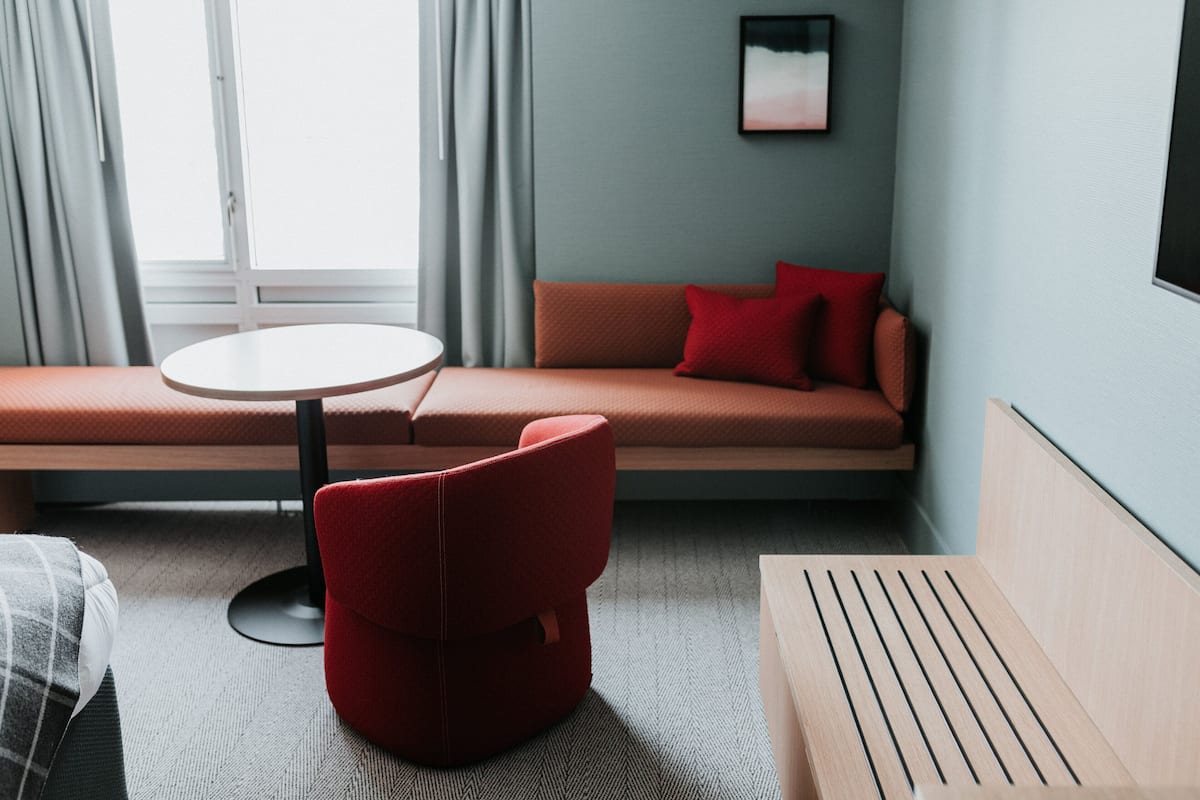 A modern hotel room with a sofa, a red chair, a round table, and a window with curtains.