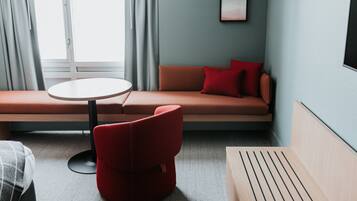 A modern hotel room with a sofa, a red chair, a round table, and a window with curtains.