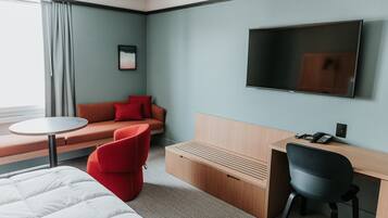 Ett hotellrum med en säng, ett skrivbord, en stol, en soffa, en tv och ett fönster med gardiner.
