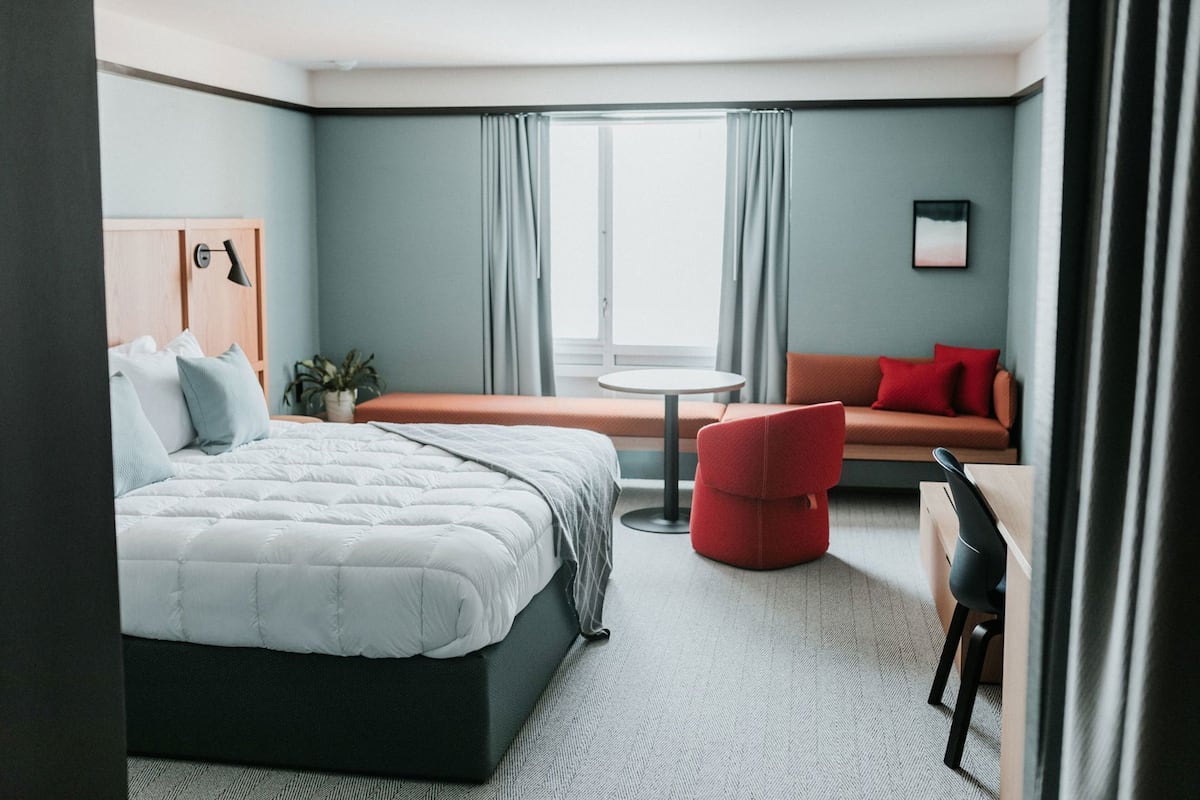 A modern hotel room with a bed, a red armchair, a small table, and a sofa.