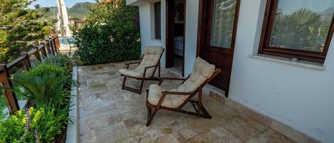 A balcony with two lounge chairs, a stone tiled floor, and a view of the surrounding greenery and mountains.