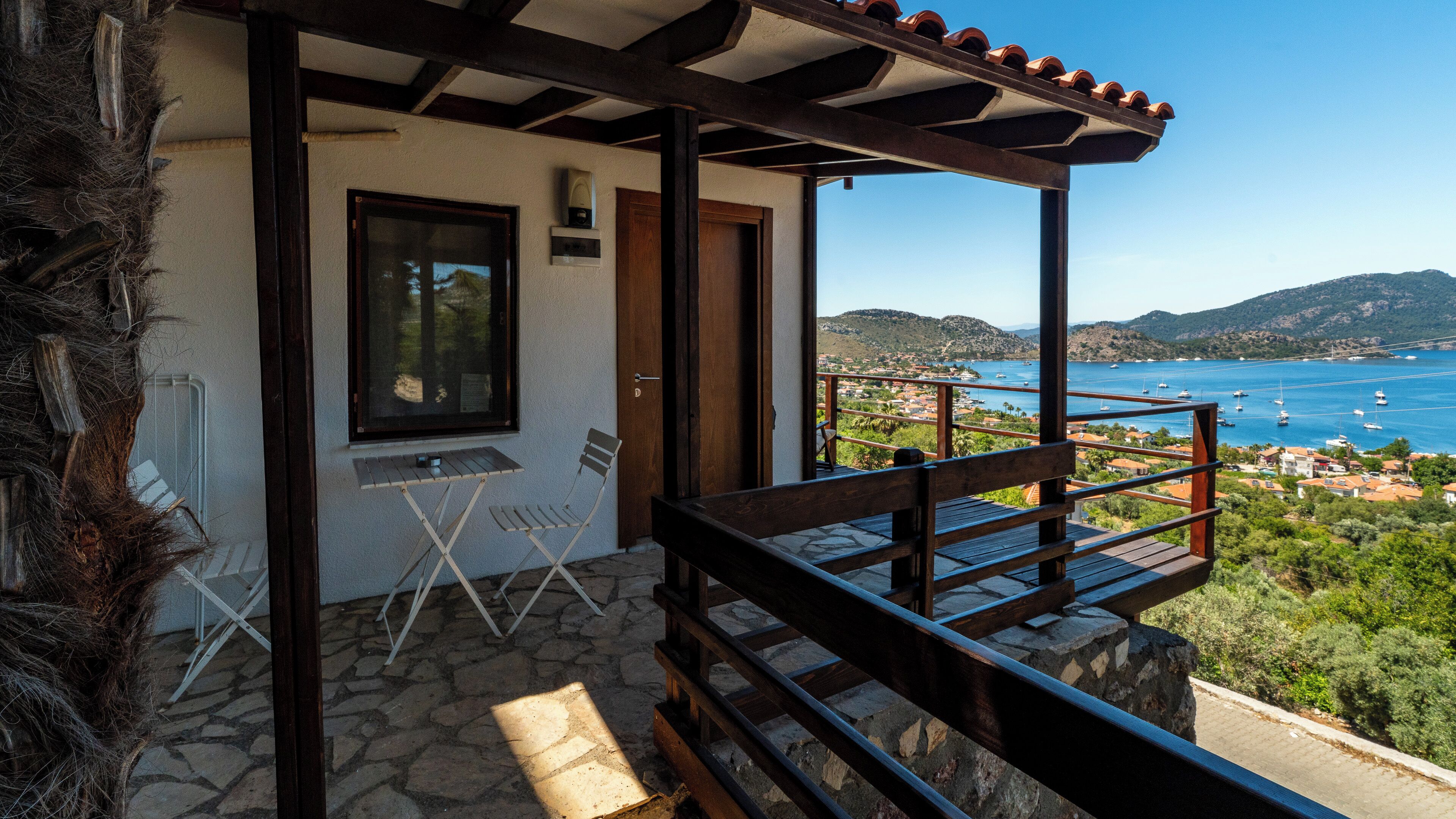 A balcony with a stone floor, a wooden bench, and a view of the sea and hills.