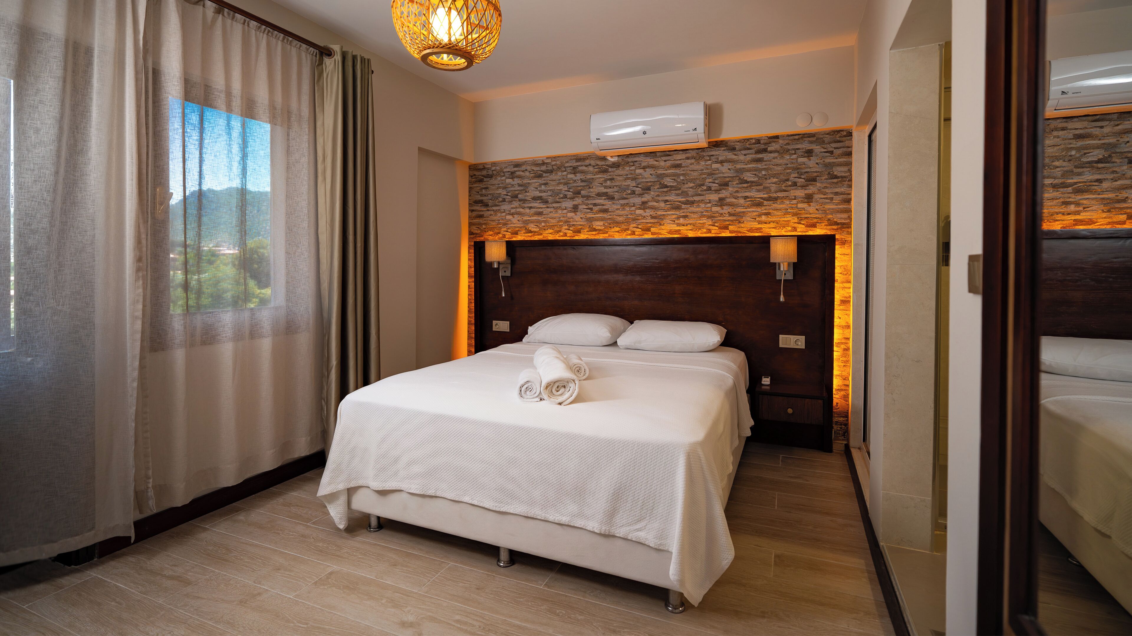 A hotel room with a bed, a window with curtains, a wall-mounted air conditioner, and a wooden headboard.