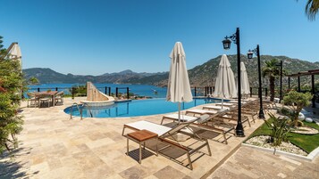 Ένας χώρος με πισίνα, ξαπλώστρες, ομπρέλες και θέα στη θάλασσα και τα βουνά.
